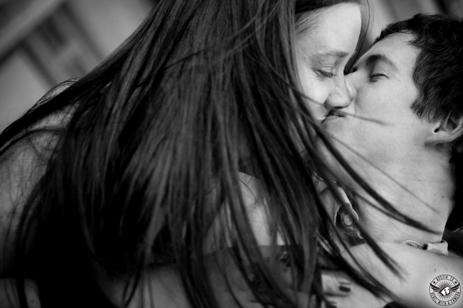 Windswept, brown hair girl with eyes closed  kisses brunette guy with eyes closed wearing a light button up dress shirt in at the Texas Capital Grounds in this powerful engagement image in Austin, Texas